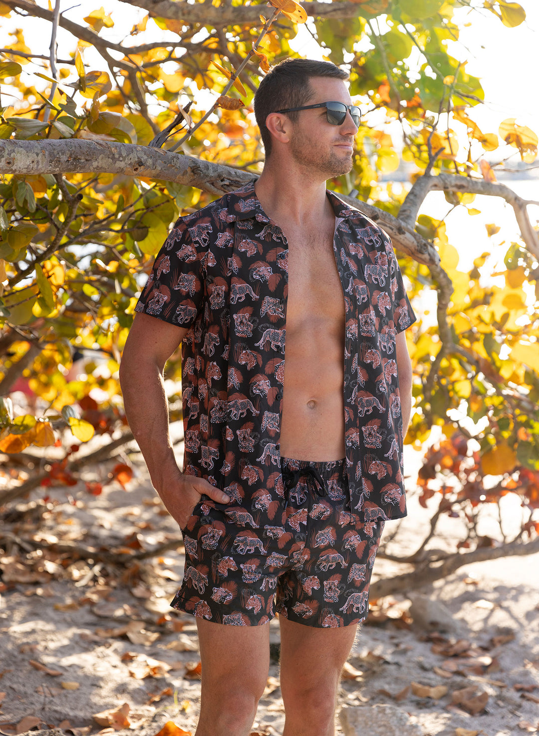 A man standing on a beach wearing a black button-up shirt with a cheetah and leopard print and matching shorts. Salty Mermaid brand.