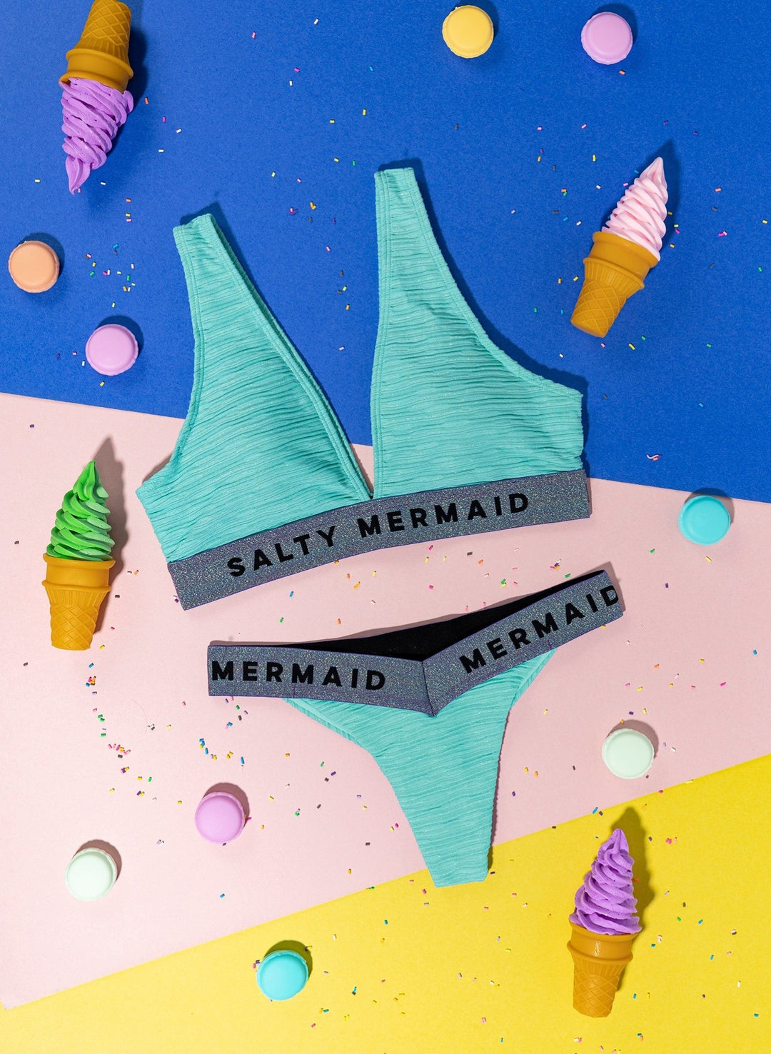Salty Mermaid seafoam green banded bikini set flat lay with elastic logo bands, sporty women’s swimwear bralette and cheeky bottoms on a multicolored backdrop with ice cream and sprinkles