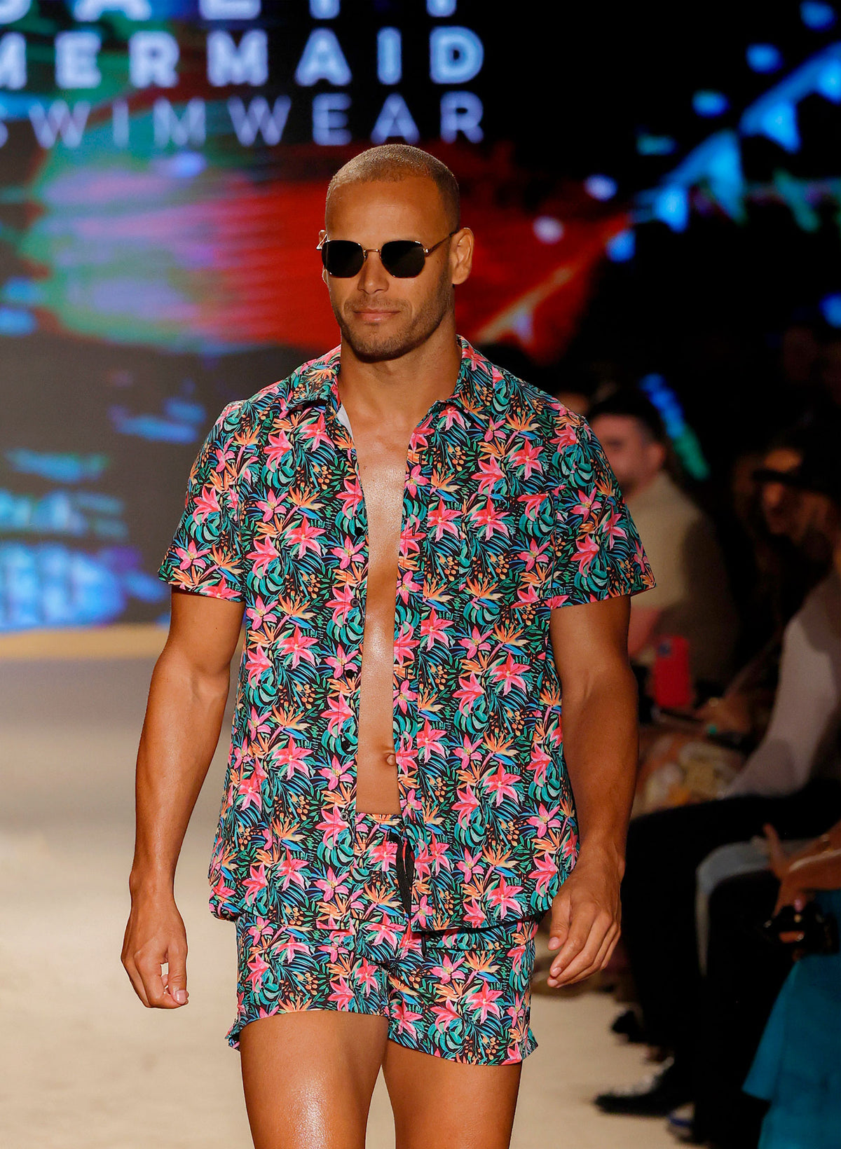 Men’s Island Bloom swim trunks featuring vibrant tropical floral print, front and back views with braided drawstring waistband, zipper pocket, and mesh liner with hidden pocket. Styled with matching shirt for coordinated beachwear. Perfect for couples matching swimwear in the Island Bloom collection by Salty Mermaid. Men’s Island Bloom button-up shirt in tropical floral print, shown in front and back views. Styled open over matching swim trunks for a coordinated resort outfit. Features lightweight fabric,