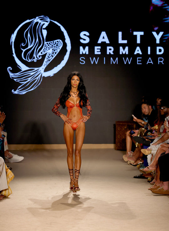 Model walking at Miami Swim Week Paraiso show wearing a Salty Mermaid red one-piece swimwear with brand logo in the background.