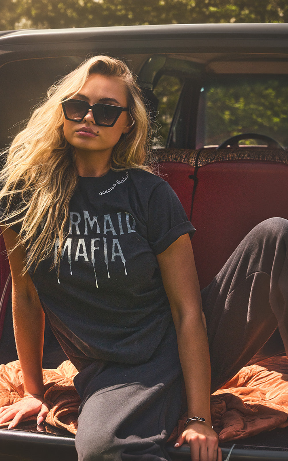 A woman wearing a black graphic t-shirt with 'MERMAID MAFIA' printed in white dripping text, sitting in the back of a vintage car.