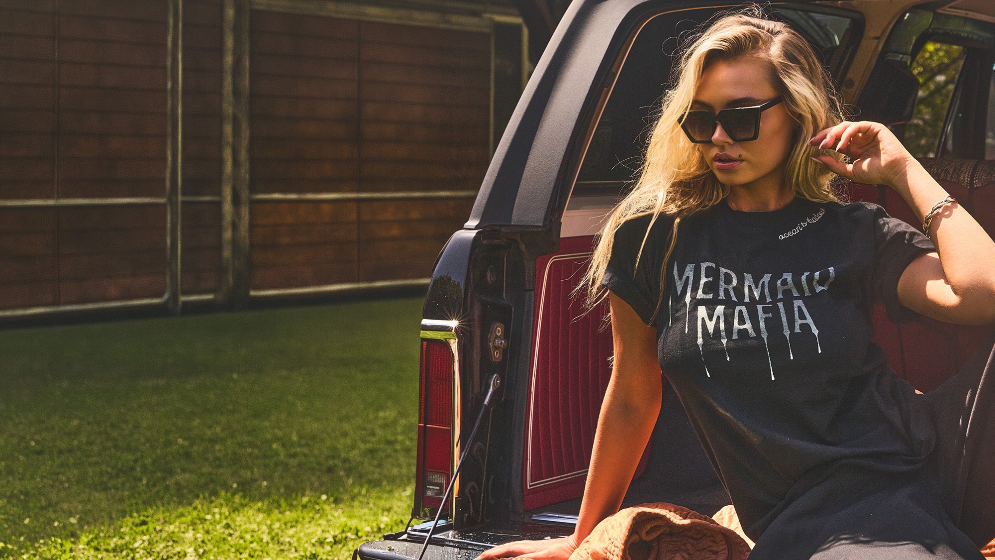 A woman wearing black sunglasses and a black graphic t-shirt with 'MERMAID MAFIA' printed in white dripping letters, sitting in the open trunk of a vehicle outdoors.