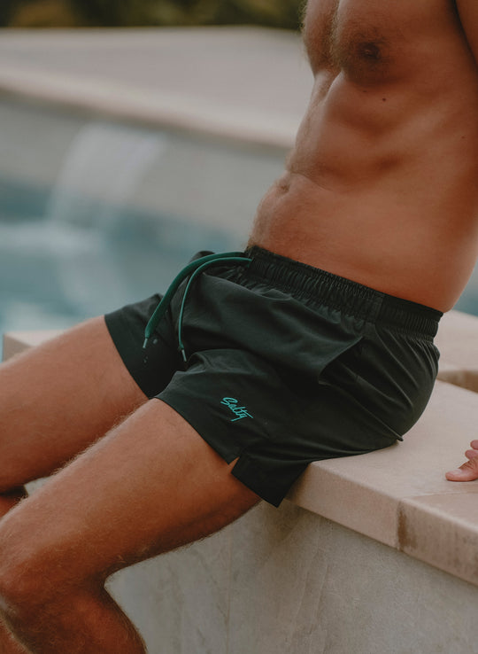 Salty Mermaid men’s black 5-inch swim trunks with Evergreen drawstring and logo embroidery styled by the pool.