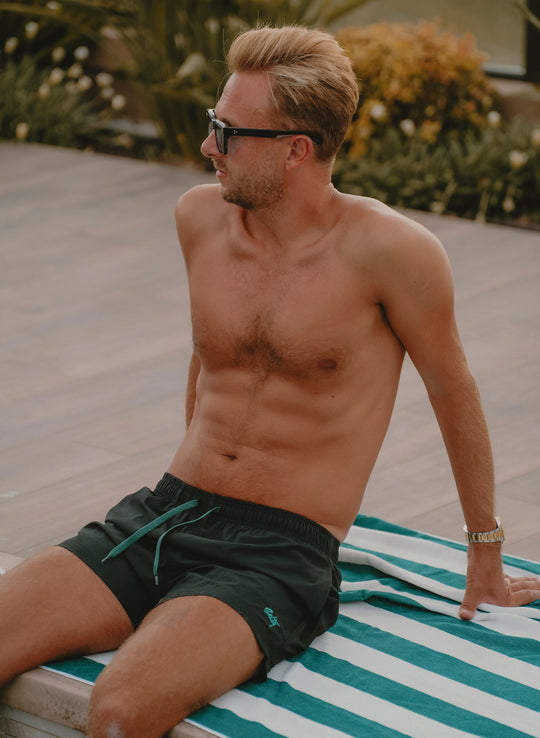 Model wearing Salty Mermaid men’s black 5-inch swim trunks with Evergreen accents while sitting poolside.