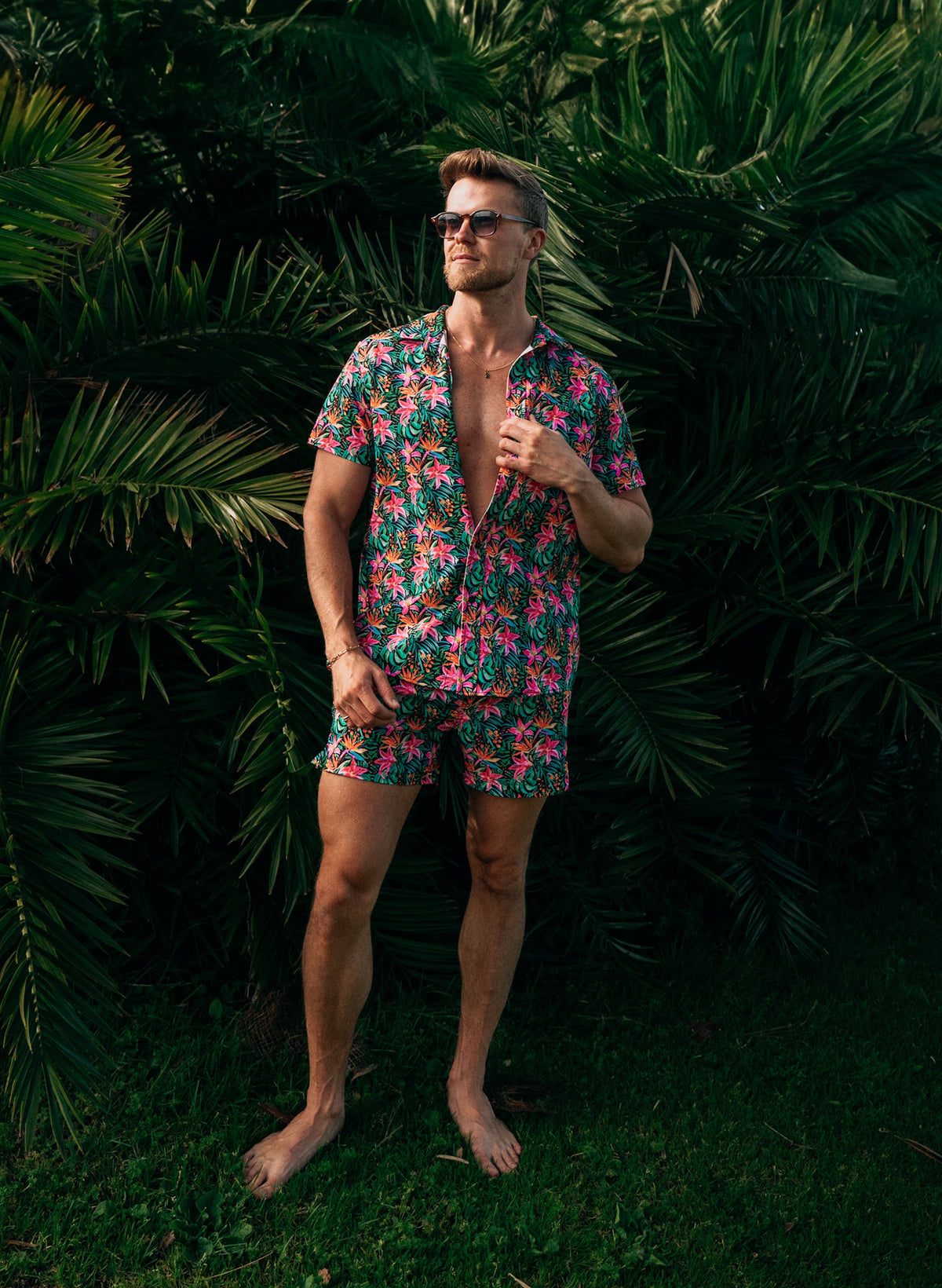 Men’s Island Bloom swim trunks featuring vibrant tropical floral print, front and back views with braided drawstring waistband, zipper pocket, and mesh liner with hidden pocket. Styled with matching shirt for coordinated beachwear. Perfect for couples matching swimwear in the Island Bloom collection by Salty Mermaid. Men’s Island Bloom button-up shirt in tropical floral print, shown in front and back views. Styled open over matching swim trunks for a coordinated resort outfit. Features lightweight fabric,
