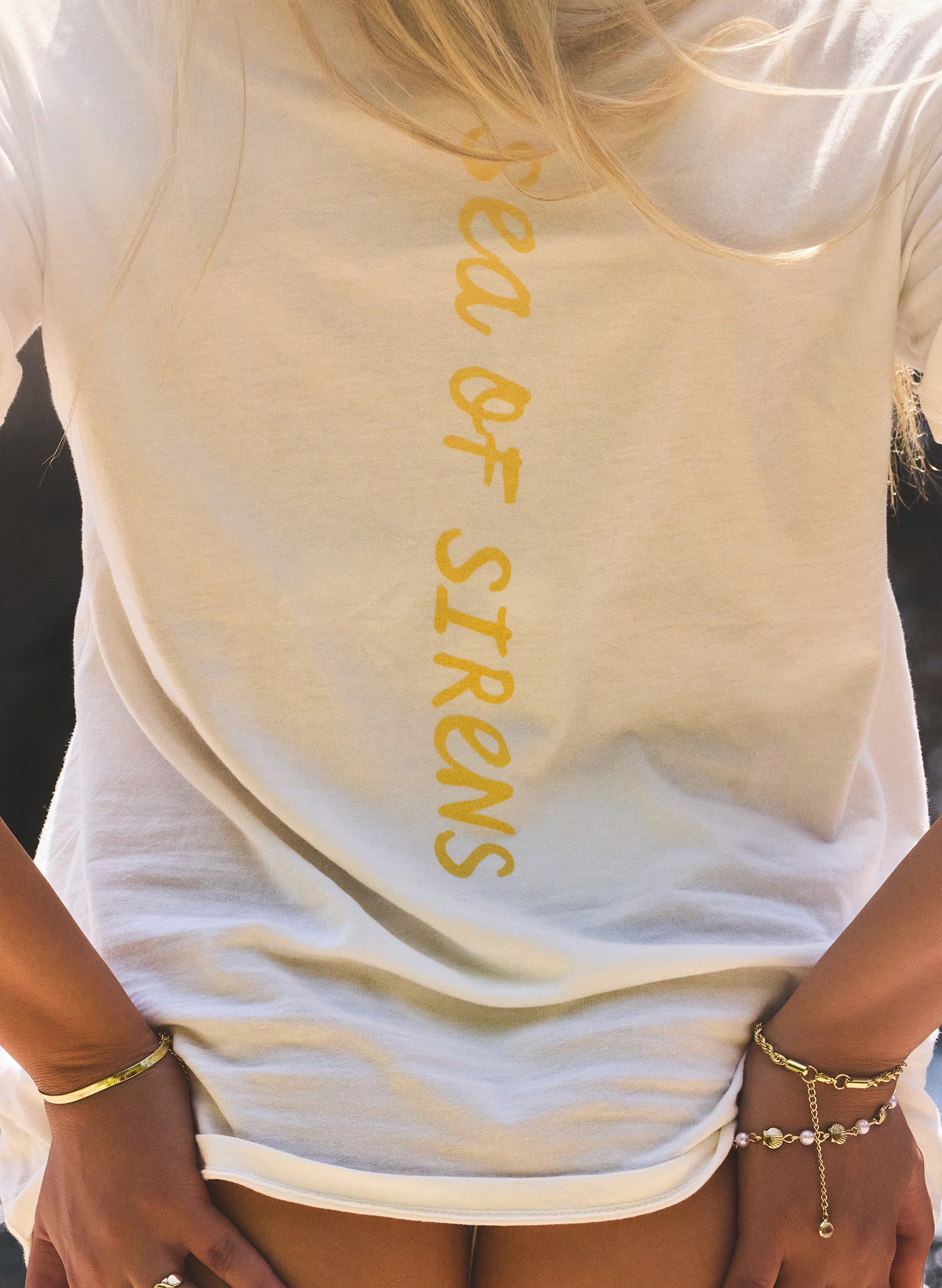Close-up back view of the white Salty Mermaid Shell Kissed unisex T-shirt featuring vertical gold “Sea of Sirens” lettering, styled as an oversized beach cover-up with gold jewelry.