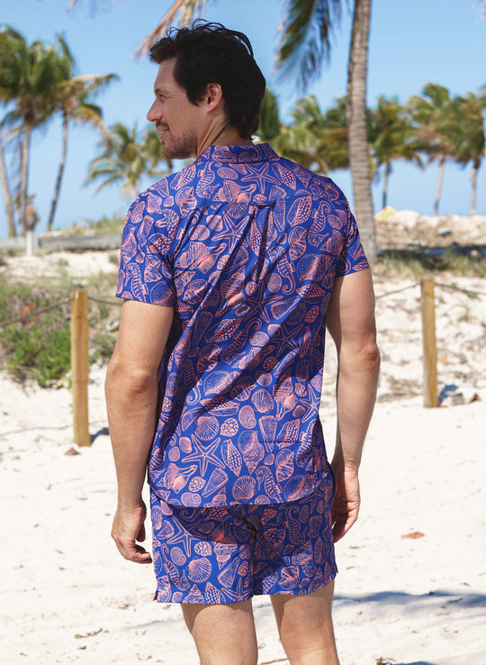 Back view of a male model wearing the matching Salty Mermaid Neon Drift button-down shirt and swim trunks, designed in a bold electric-blue print with coral-pink and purple illustrated seashells, on a palm-lined beach.