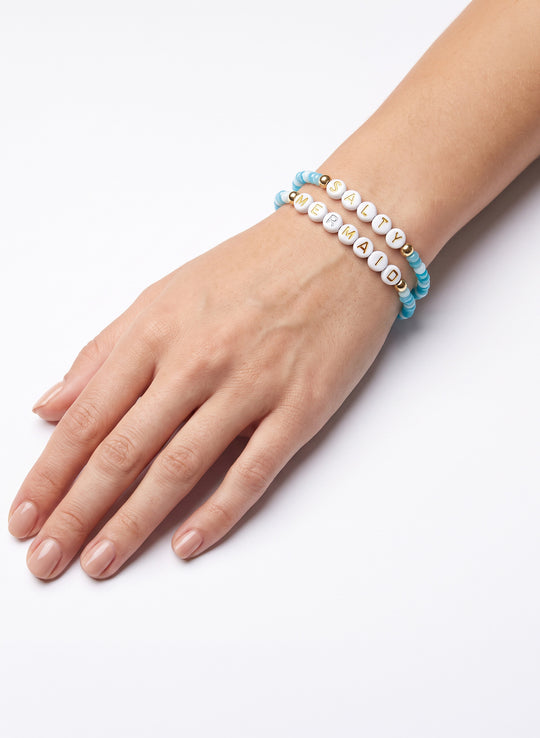 A close-up of a person's hand and wrist wearing two beaded bracelets on a clean white background. One bracelet is light blue with white beads, and the other is white with gold lettering spelling 'JENNY' and small gold beads.