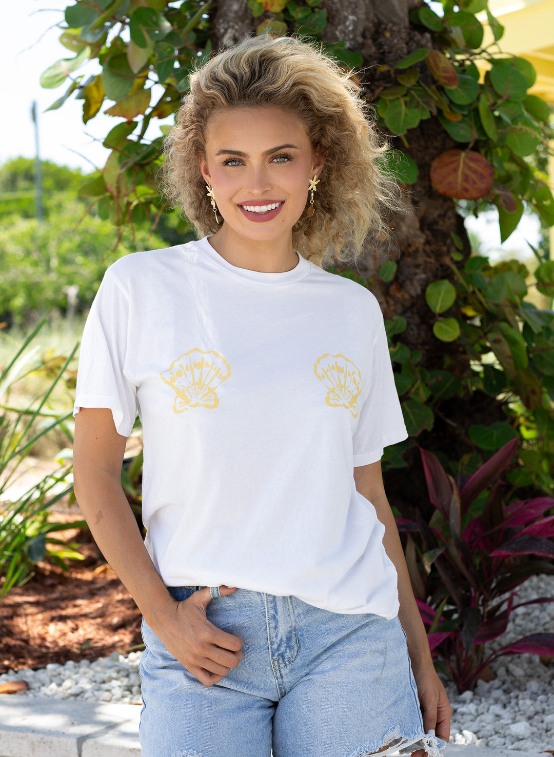 Female model wearing the Salty Mermaid Shell Kissed white T-shirt with twin gold seashell graphics, styled in a relaxed fit with light-wash distressed denim shorts near the beach.