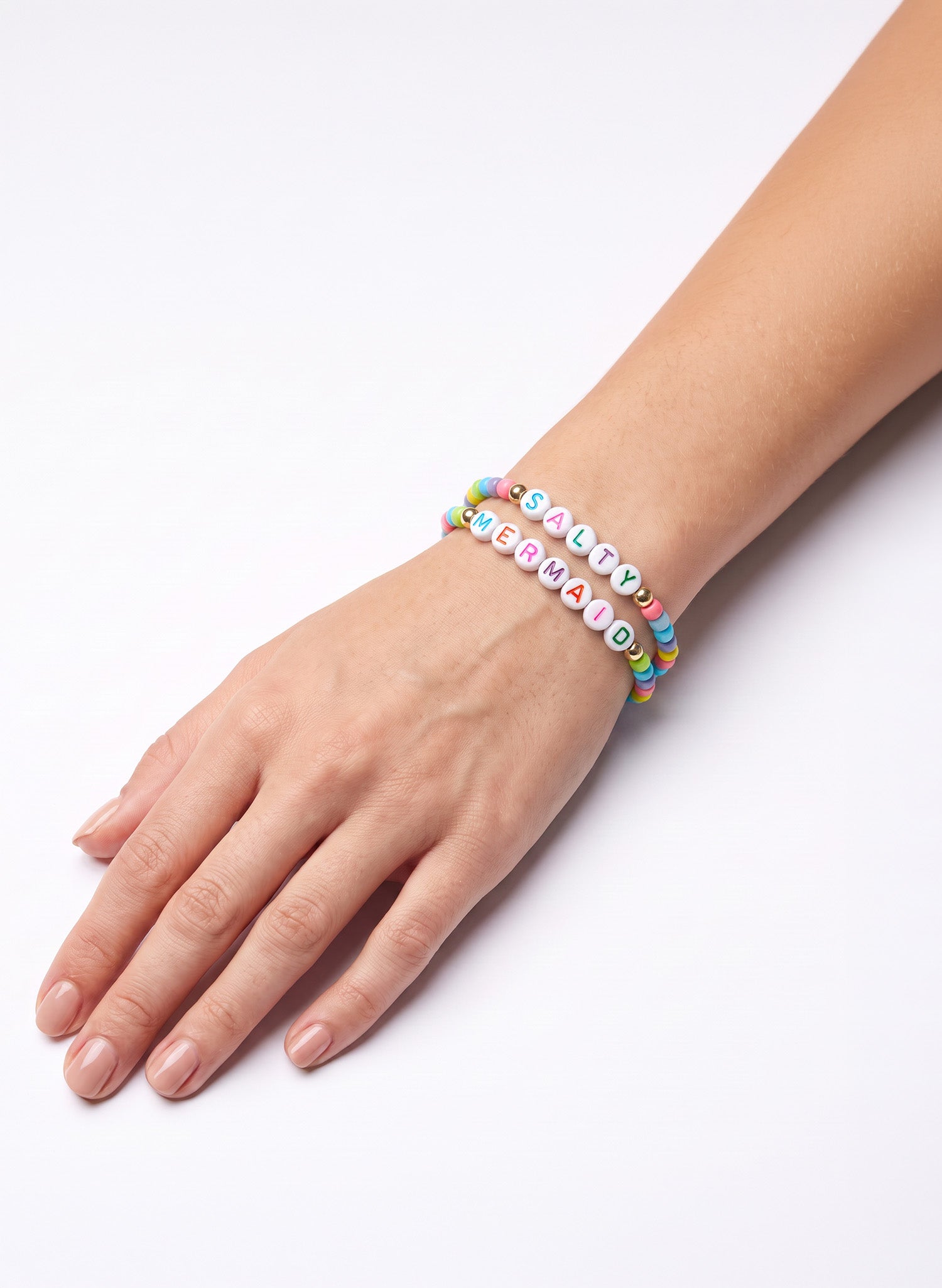 A close-up of a person's hand and wrist wearing two colorful beaded bracelets on a plain white background. One bracelet features pastel rainbow-colored beads, while the other has white letter beads spelling 'SALTY MERMAID'.