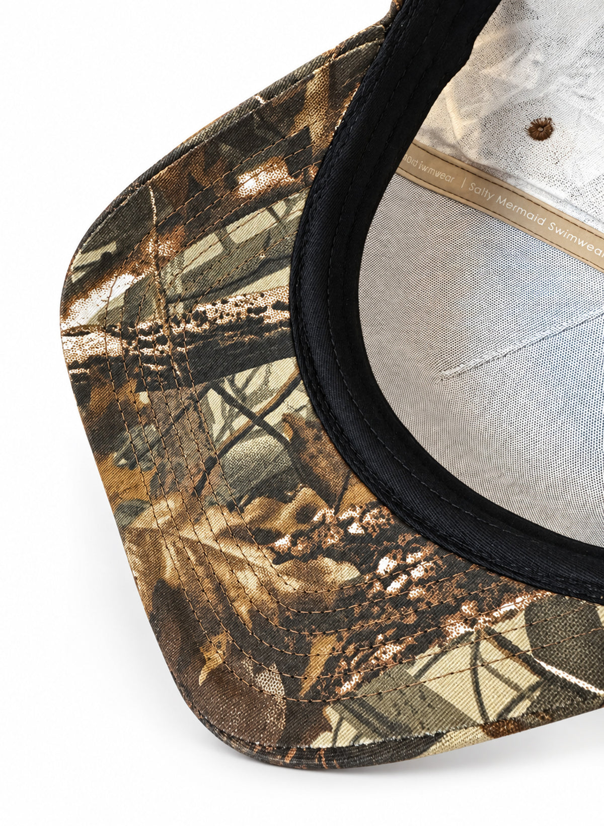 Close-up of the Dark Woodland camo hat’s patterned brim underside, black sweatband, and interior seam tape on white.

