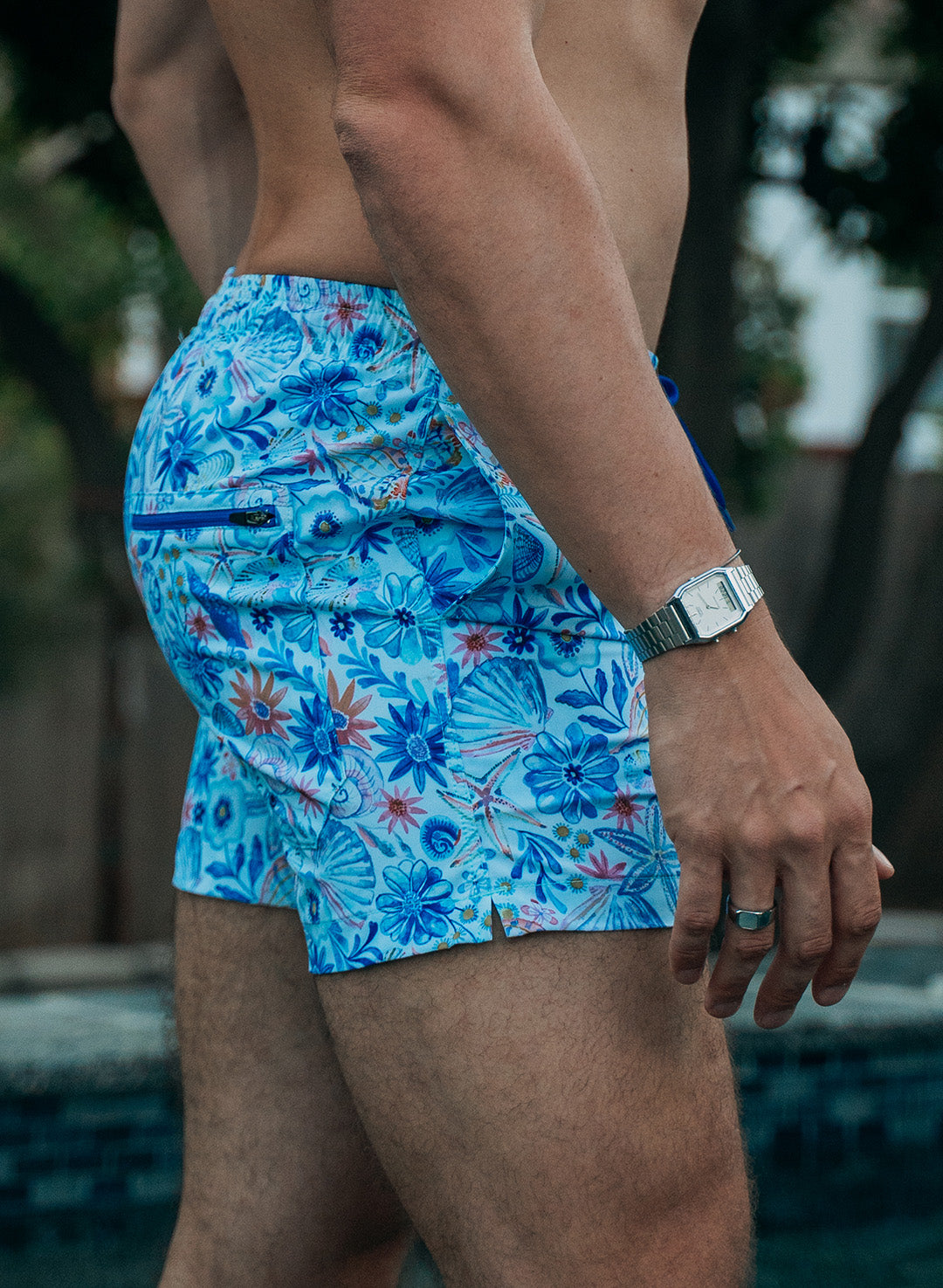 Salty Mermaid men’s swim trunks from the Nautical Garden collection with tropical floral and seashell motif in a hand-drawn style, featuring a zippered back pocket and open side slits.