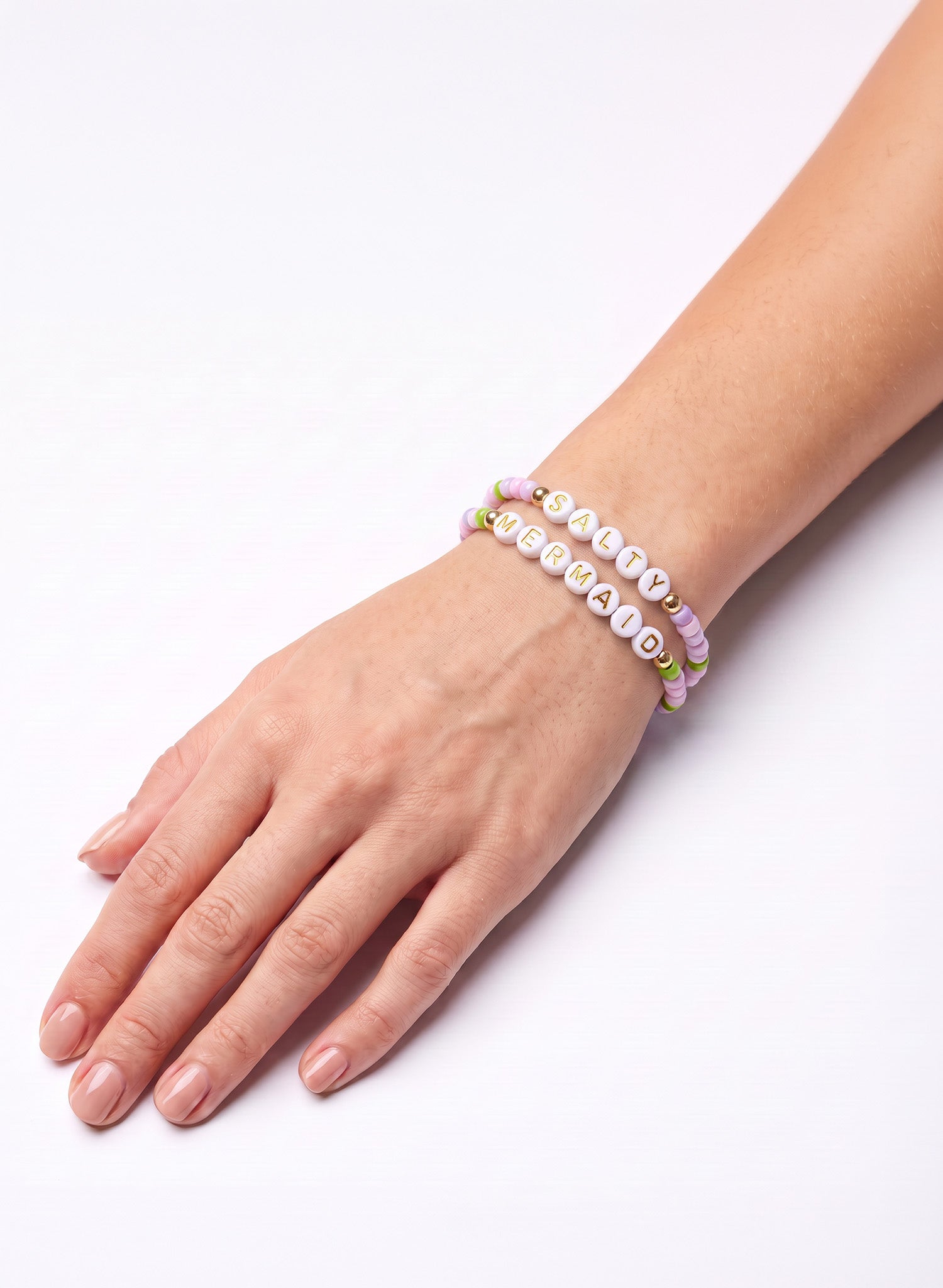 A close-up of a person's hand and wrist wearing two beaded bracelets on a plain white background. One bracelet features white letter beads spelling 'Salty', and the other has pastel pink, purple, and green beads.