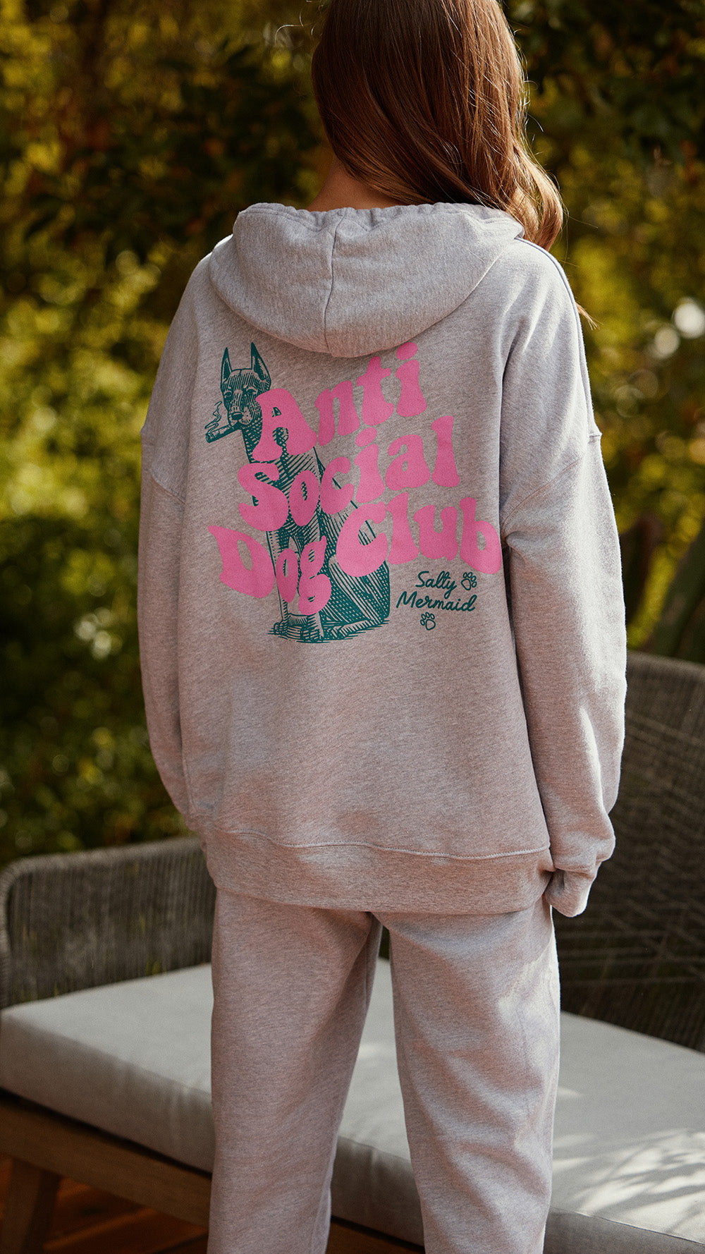 A person viewed from behind wearing a light heather gray hoodie and matching sweatpants, with a large pink and teal graphic on the back reading 'Anti Social dog Club' and 'Sally's Mermaid'. The setting is an outdoor patio with blurred greenery in the background