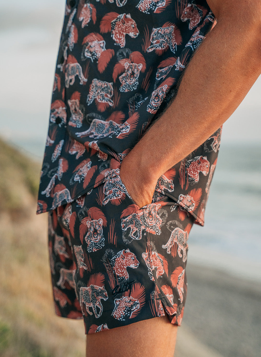 Close-up of a man wearing Salty Mermaid black swim trunks with red leopard and palm leaf print and matching shirt.