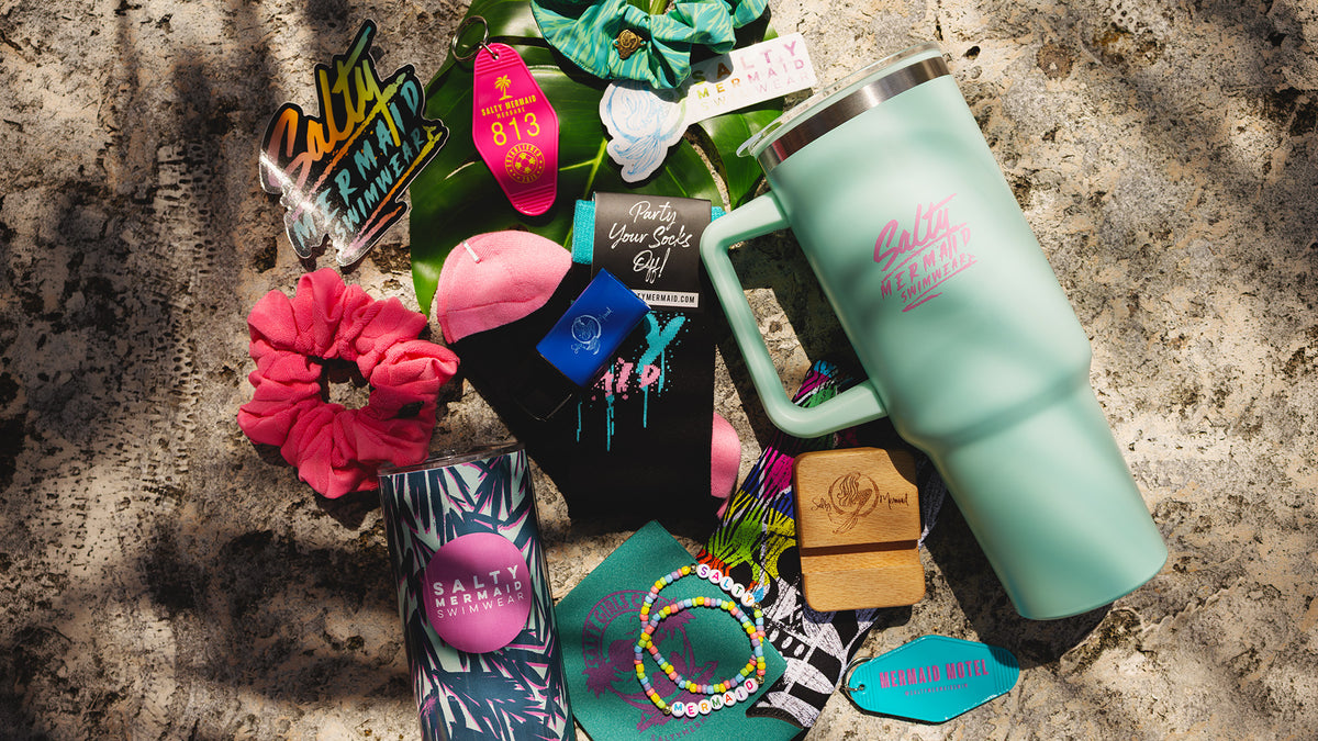A styled flat-lay of various branded salty mermaid mystery accessory box on a textured concrete surface with dappled sunlight. The collection includes a large mint green insulated travel mug with a pink scrunchie, a black sock with colorful graphics, a wooden stamp, a bamboo box, and other matching accessories.