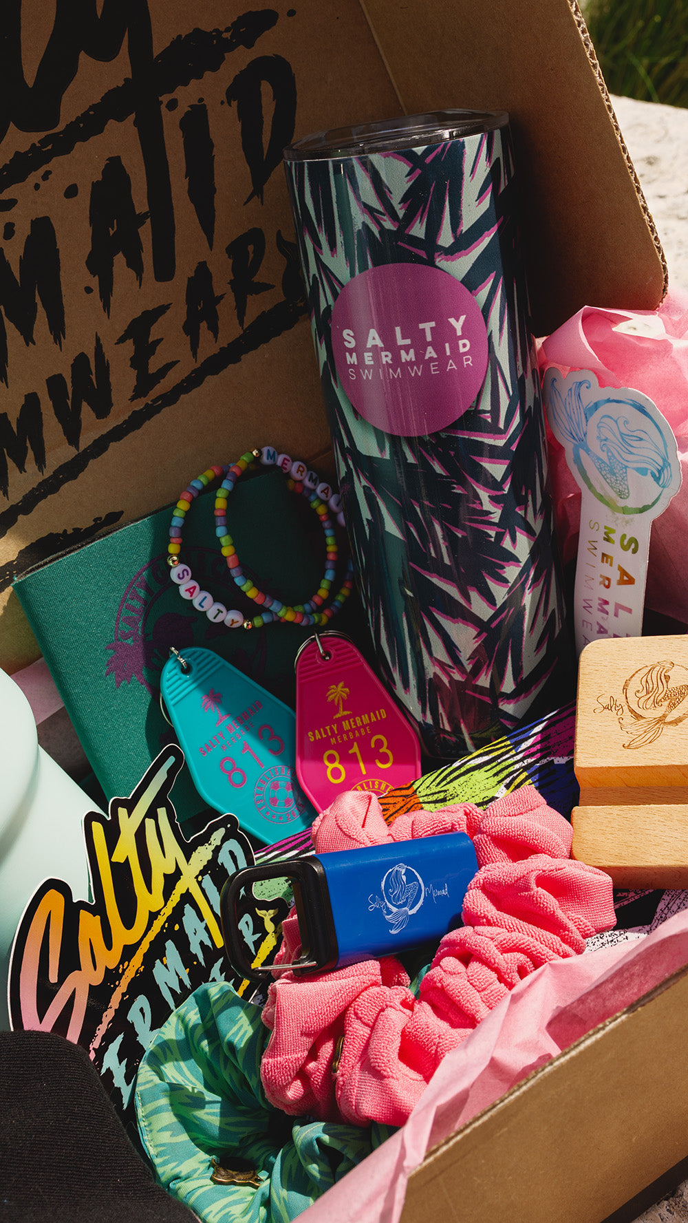 A curated gift box filled with branded Salty Mermaids products, including a tumbler with a palm tree pattern, scrunchies, a beaded bracelet, a small blue item, and pink accessories, arranged in a cardboard box.