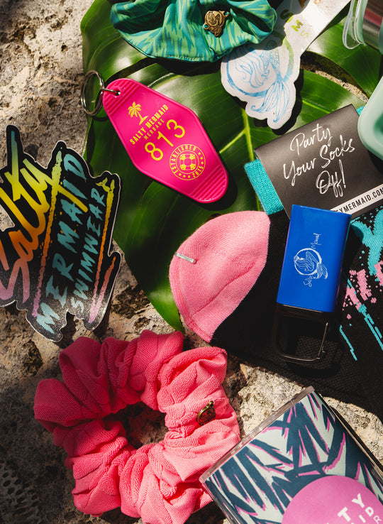 Salty Mermaid accessories with a pink motel keychain, logo stickers, pink and green scrunchies, black socks and palm-print tumbler.