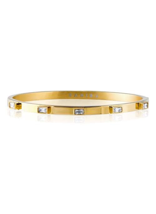 A gold sahira diamond bangle bracelet with rectangular clear gemstones set along the band, shown on a plain white background with a faint reflection below. The brand name 'SABIRA' is engraved inside the bracelet.