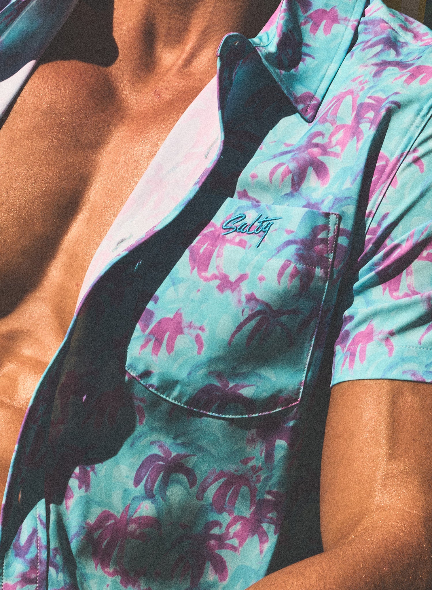 Close up of model  wearing a blue unbuttoned shirt with palm tree pattern and 'Salty' brand logo.