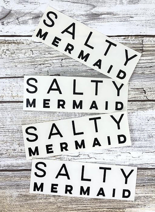 Four black vinyl stickers with the text 'SALTY MERMAID' written in white, displayed on a white wooden background.