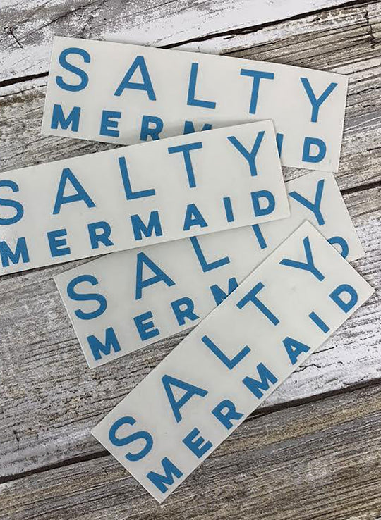 A set of four blue vinyl stickers with the text 'SALTY MERMAID' written in capital letters, placed on a white wooden surface.